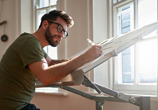 Man in glasses sitting sketching at a desk. 